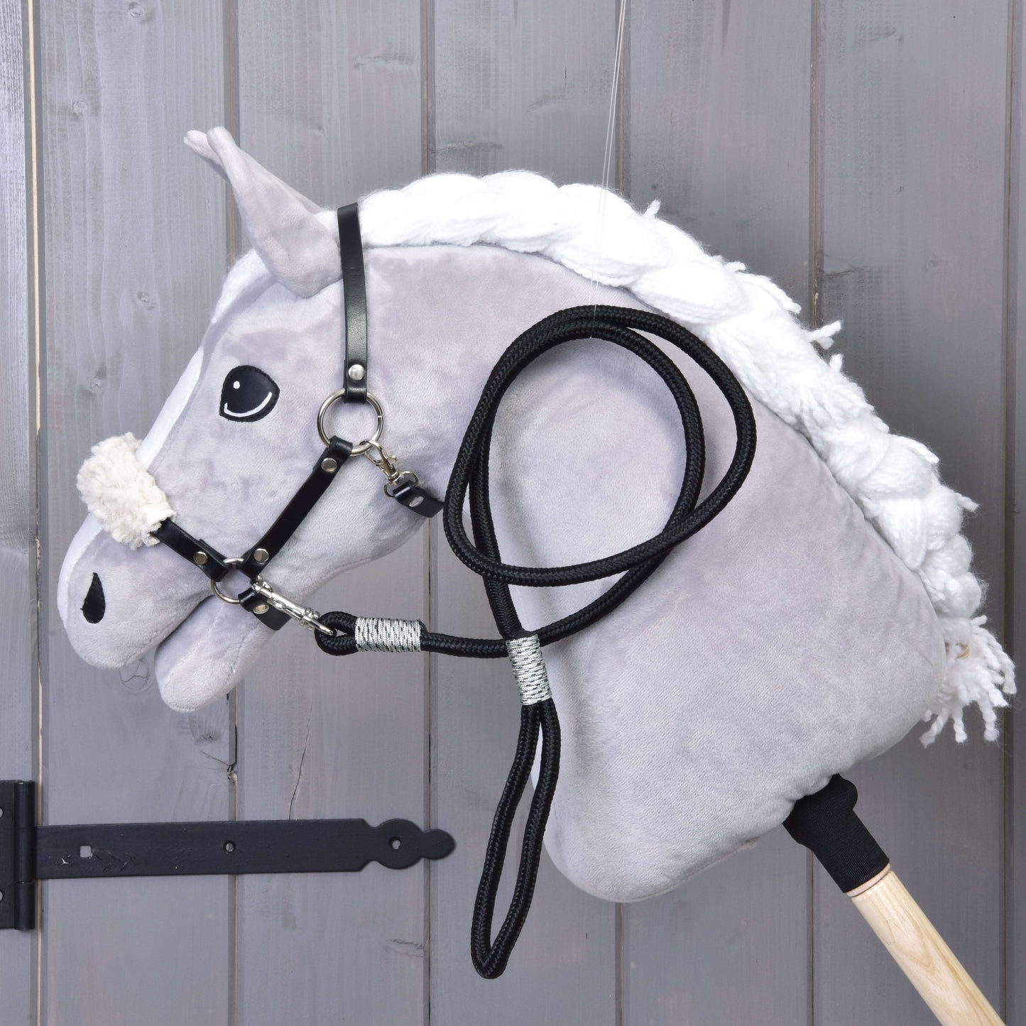 Hobby Horse LarDen Set Basic zobrazuje plyšového Hobby Horse s bílou hřívou, černou uzdečkou s otěžemi a černou ohlávkou na dřevěné tyči, opřeného o šedé dřevěné panely.