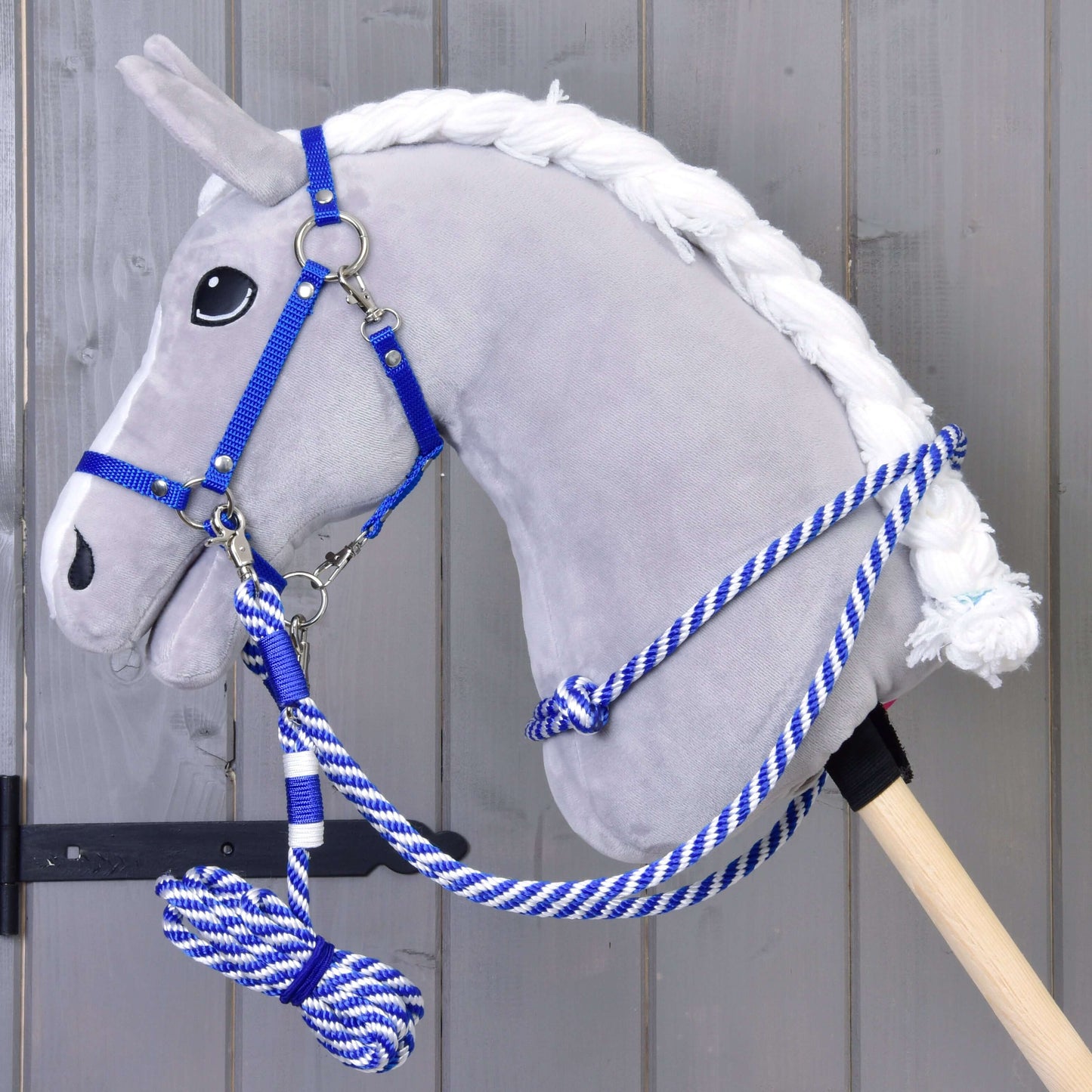 Plyšový šedý Hobby Horse od Hobby Horse LarDen se sadou Set Start modrý (ohlávka + vodítko + otěže + nákrčník), s modro-bílým vodítkem na dřevěné tyči, stojí před šedou dřevěnou stěnou.