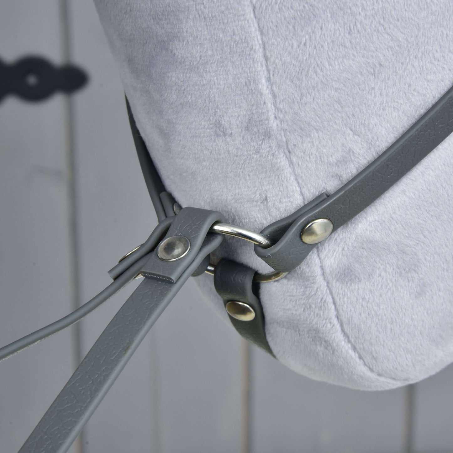 Martingale tmavě šedý od Hobby Horse LarDen na světle šedé plyšové koňské hlavě, s šedými koženými řemínky a kovovými kroužky na rozmazaném dřevěném pozadí.