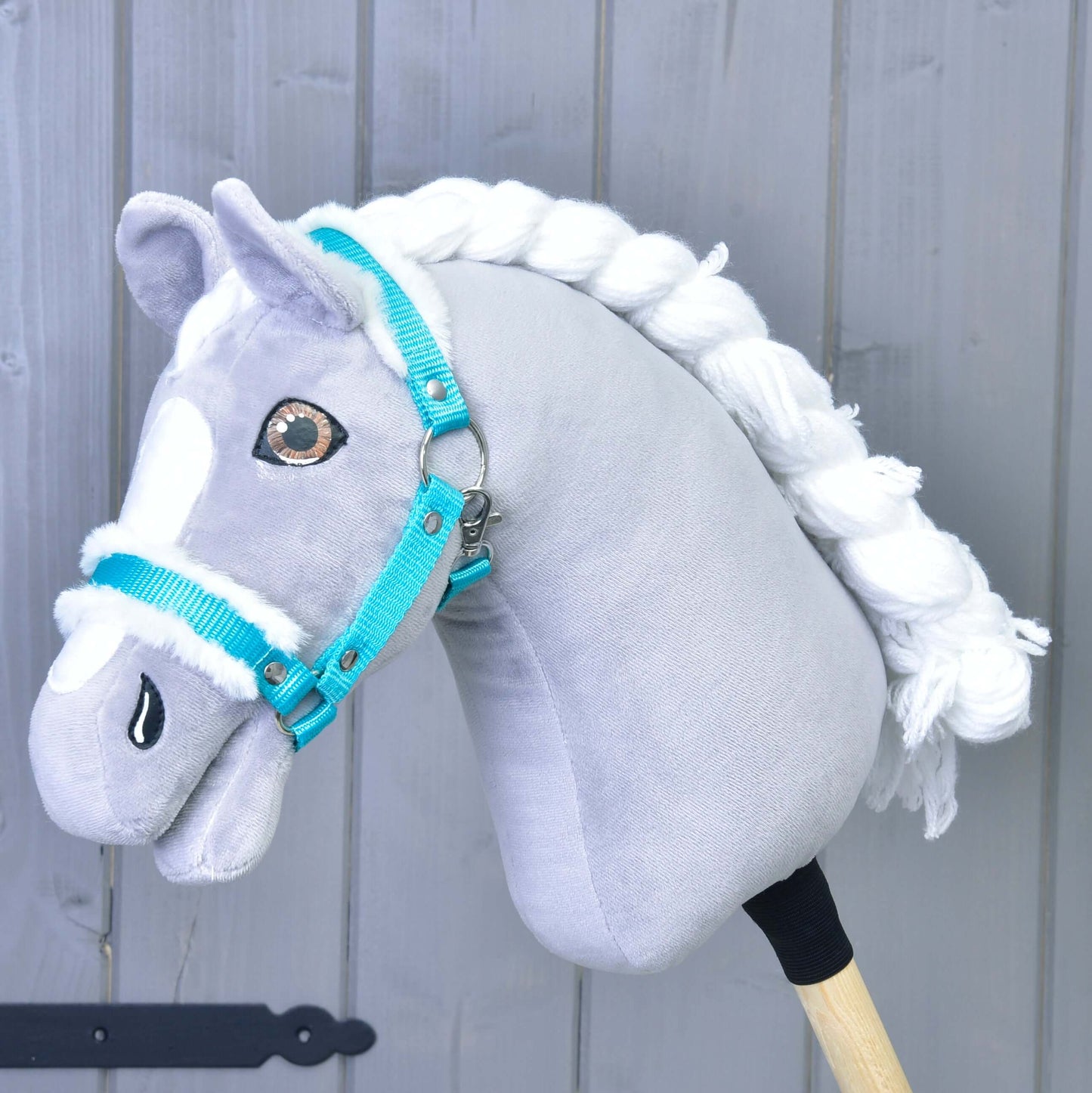 Šedo-bílý Hobby Horse se zapletenou bílou hřívou má na sobě Hobby Horse LarDen „Ohlávka tyrkys s beránkem“, perfektní doplněk, na světle šedém dřevěném pozadí.