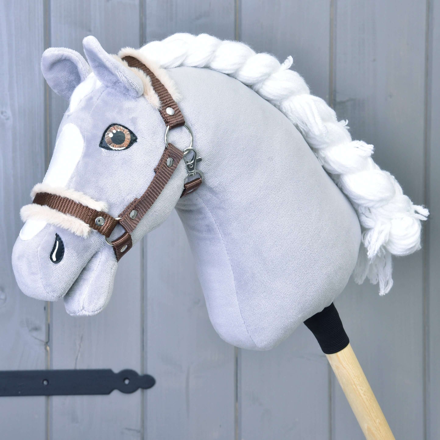 Detailní záběr na Hobby Horse LarDen "Ohlávka hnědá s beránkem" – šedý Hobby Horse s bílou hřívou a hnědou ohlávkou – na dřevěné tyči, před šedým dřevěným pozadím.