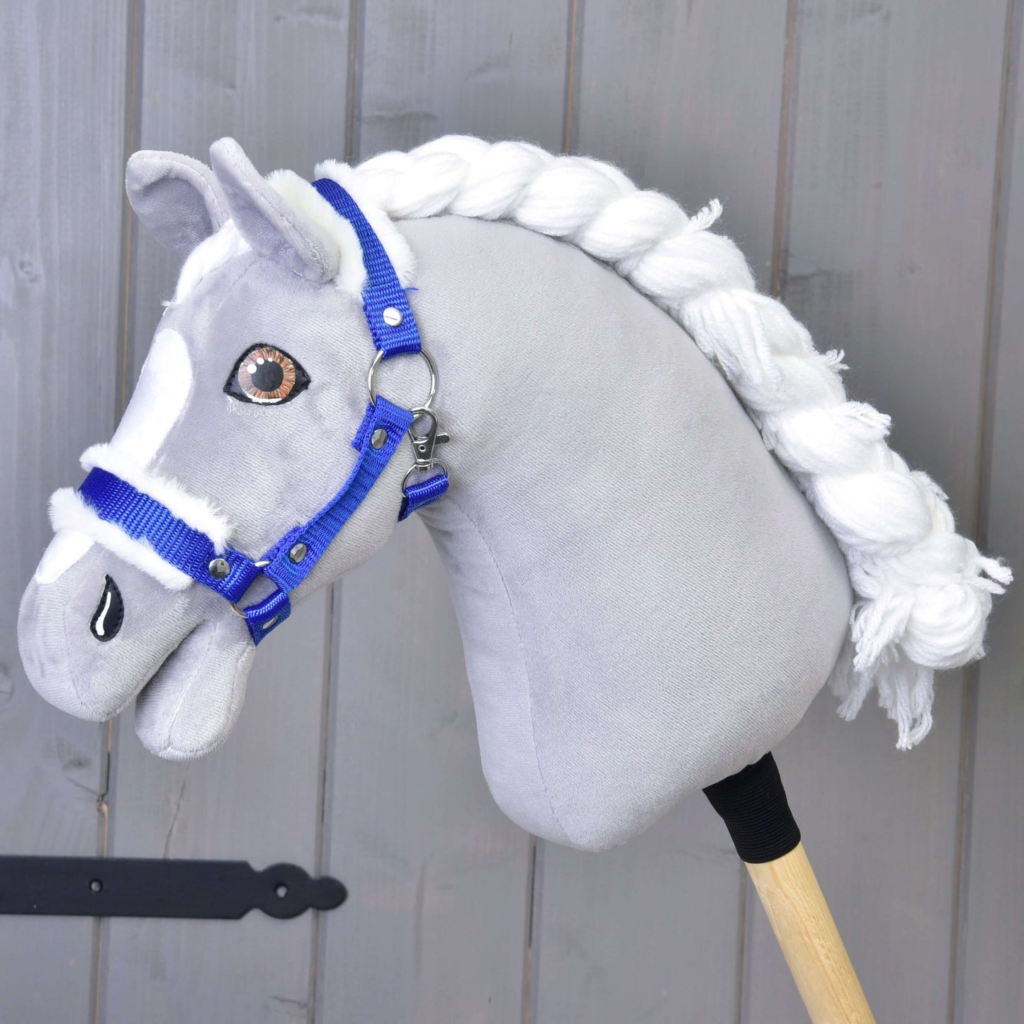 Šedý látkový Hobby Horse s bílou přízovou hřívou a ohlávkou Hobby Horse LarDen Ohlávka modrá s beránkem stojí před světle šedou dřevěnou stěnou.