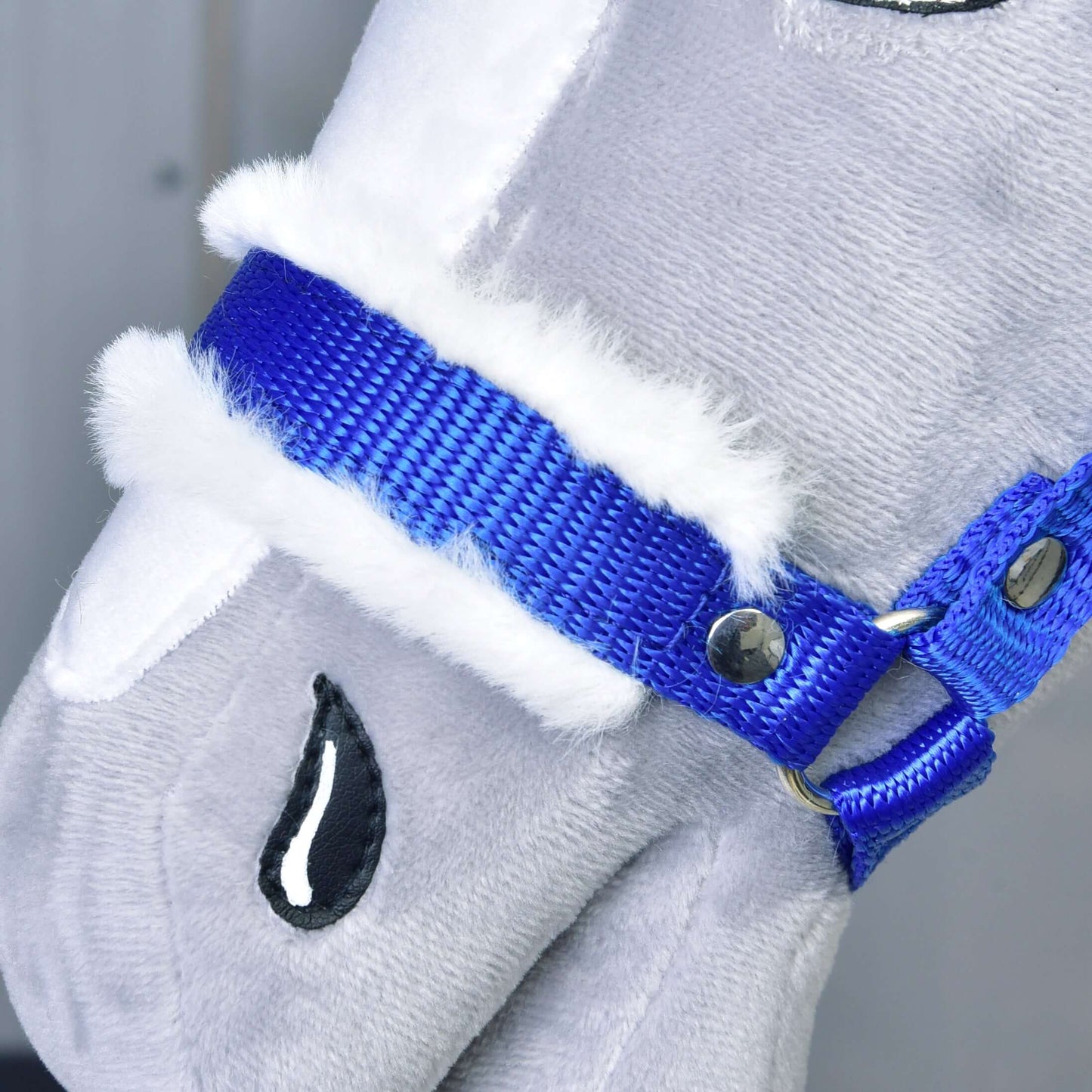 Detailní záběr na obličej plyšového koně Hobby Horse LarDen s Ohlávkou modrou s beránkem. Mezi prvky patří lem z bílé umělé kožešiny, vyšívané černé oči s detailem kapky na světle šedé látce.