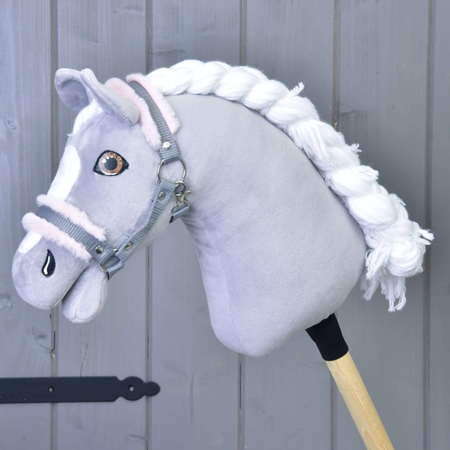 Hobby Horse LarDen "Ohlávka šedá s pink beránkem" zobrazuje šedého plyšového koně s bílou hřívou a šedou ohlávkou, připevněného k dřevěné tyči, na pozadí šedých dřevěných panelů.