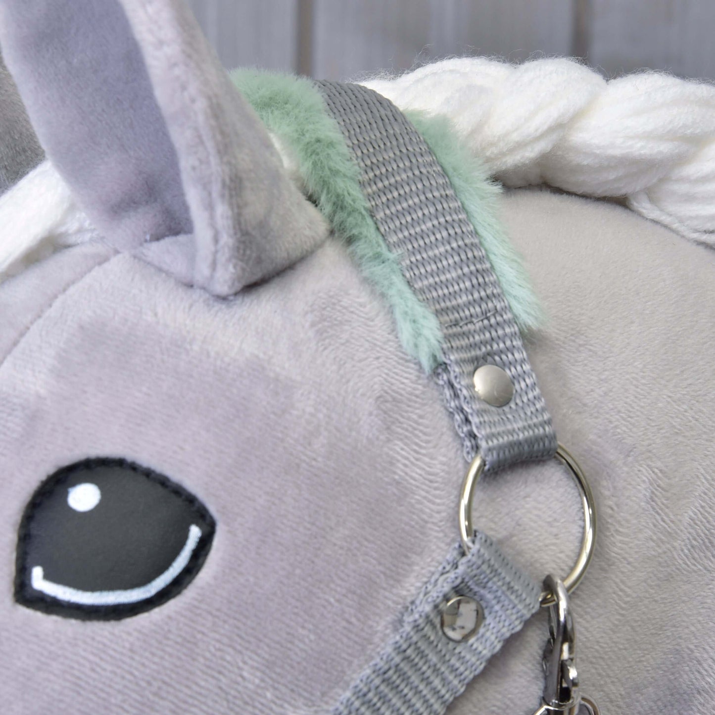 Detailní záběr šedého plyšového jednorožce s černobílým vyšívaným okem, bílou přízovou hřívou, mint zelenými akcenty a Hobby Horse LarDen "Ohlávkou šedou s mint beránkem" se stříbrným kroužkem a zapínáním.