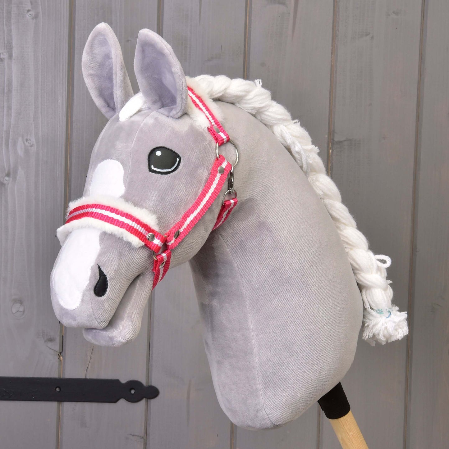 Hobby Horse LarDen "Ohlávka červený proužek s beránkem" je šedý plyšový Hobby Horse s bílou hřívou a ohlávkou s červeným proužkem na dřevěné tyči, zobrazený před šedou dřevěnou stěnou.