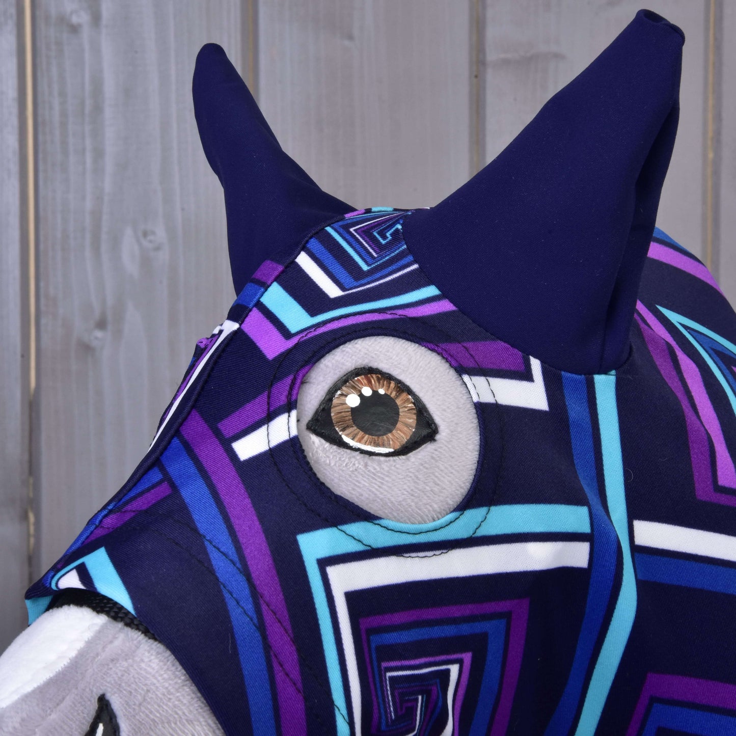 Detail Hobby Horse LarDen "Hobby Horse ochranný oblek modrý (Lycra)" ukazuje látkovou zvířecí hlavu s geometrickým fialovo-modro-bílým vzorem, velkým kulatým okem a špičatýma ušima na pozadí dřevěného panelu.