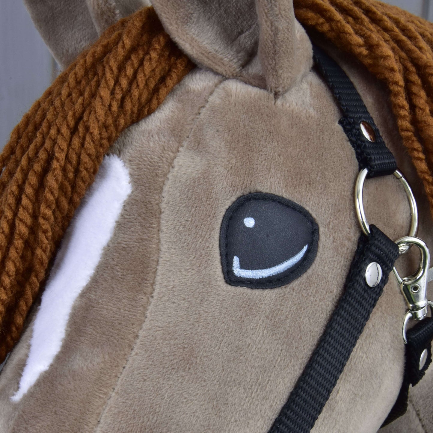 Detail Hobby Horse Oliver od Hobby Horse LarDen s černou ohlávkou a ohlávkou s beránkem zdarma, s hnědou pletenou hřívou z příze, bílou skvrnou kolem oka a našitým okem v kresleném stylu, s vzhledem roztomilého drezurního koně.