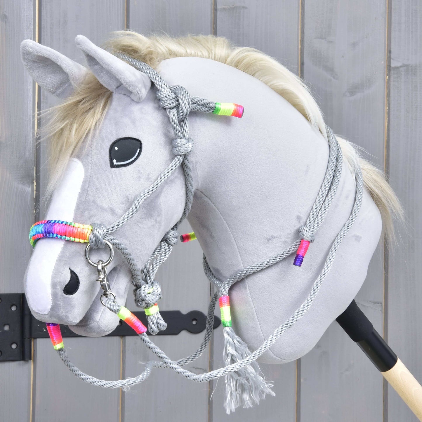 Šedý Hobby Horse s bílou lysinou, černým nosem a blond hřívou má na sobě Provazový set Manhattan od Hobby Horse LarDen se šedou provazovou ohlávkou a otěžemi s barevnými korálky, na dřevěném pozadí.