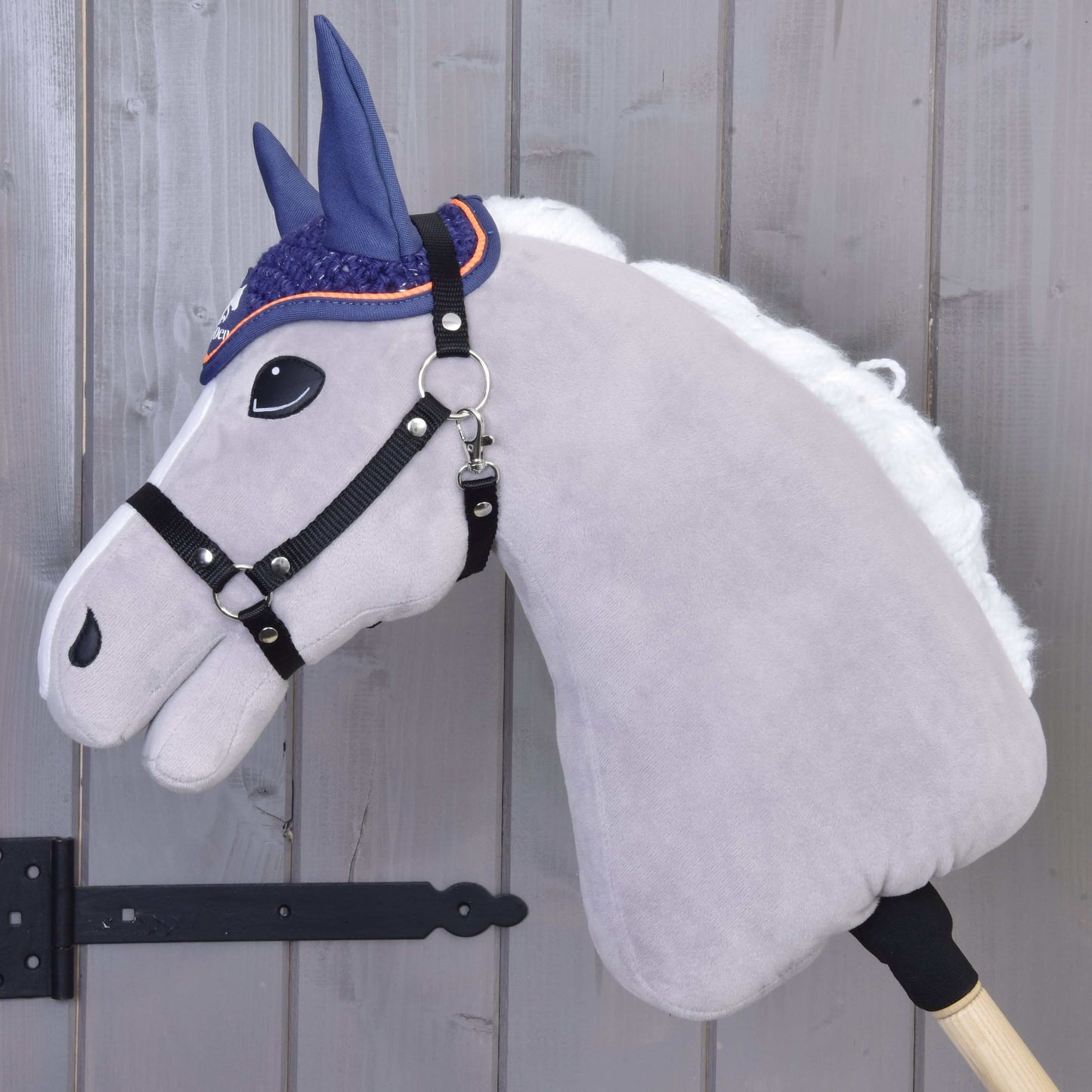 Čabraka háčkovaná Blue sport od Hobby Horse LarDen na plyšové světle šedé hlavě s bílou hřívou, černou uzdečkou a modrou háčkovanou čabrakou na dřevěné tyči, před světlou dřevěnou stěnou.