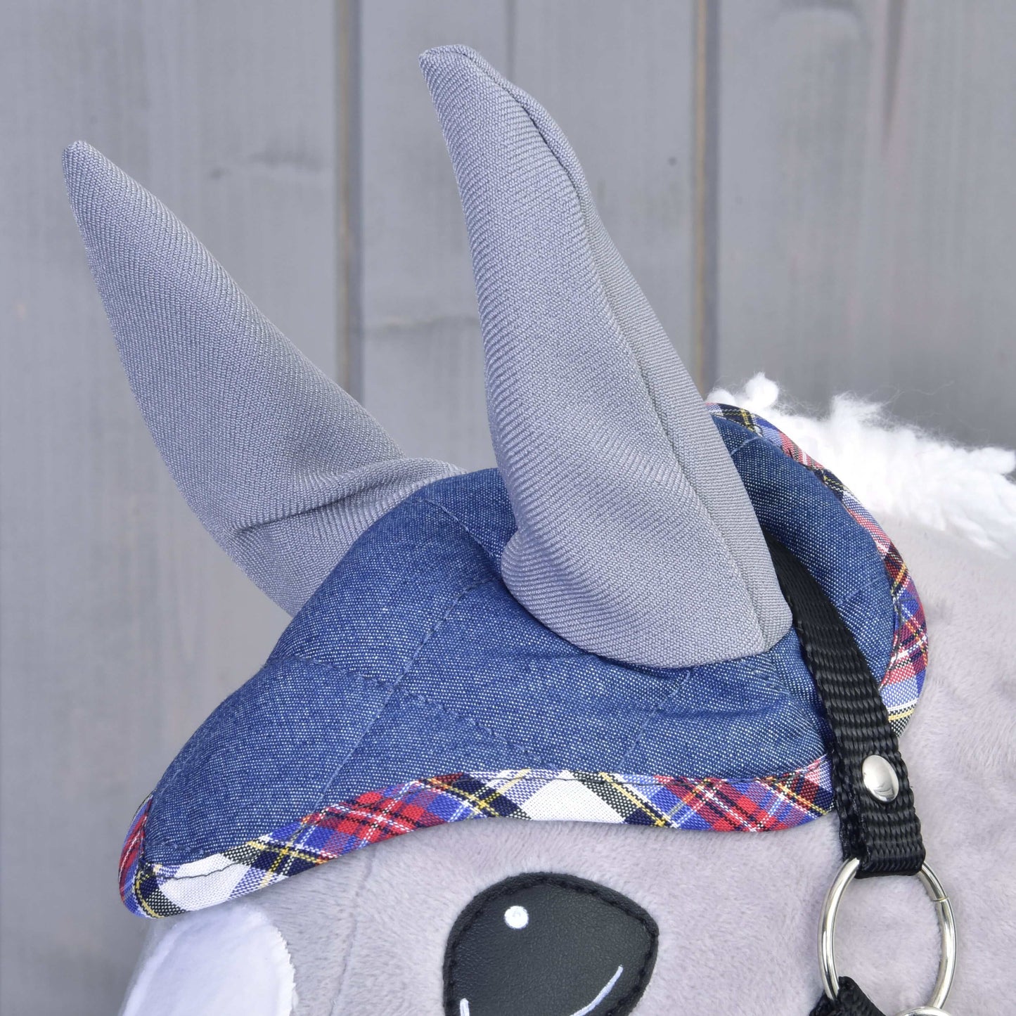 Detail plyšového zvířete Set Jeans od Hobby Horse LarDen se šedýma látkovýma ušima, modrou kostkovanou čepicí připomínající sadu čabraky, černým popruhem s kovovým kroužkem a velkým bílým a černým okem na dřevěném pozadí.
