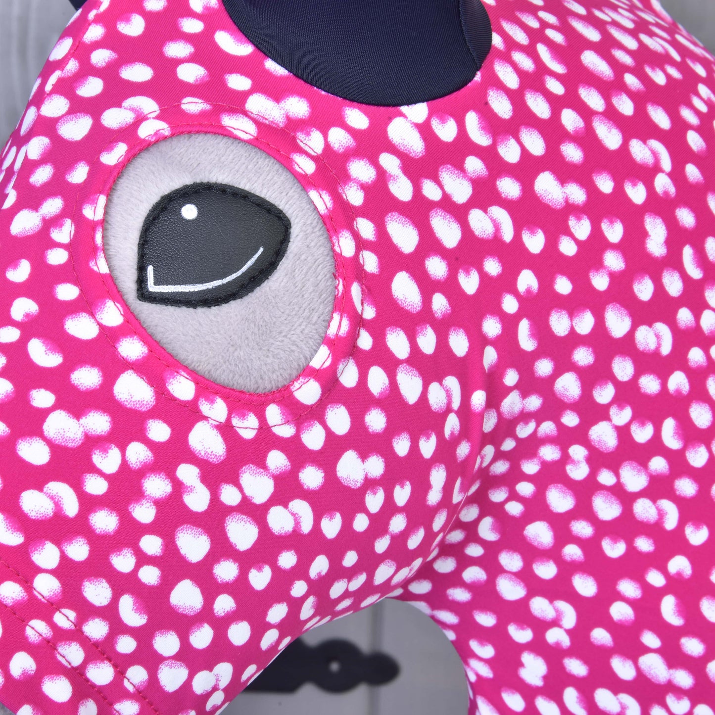Detail Hobby Horse ochranný oblek Minie (Lycra) od Hobby Horse LarDen, růžového pratelného potahu s bílými puntíky a vyšívaným šedým okem. Udržuje váš Hobby Horse čistý a stylový. Rozmazané pozadí ukazuje část šedé stěny.