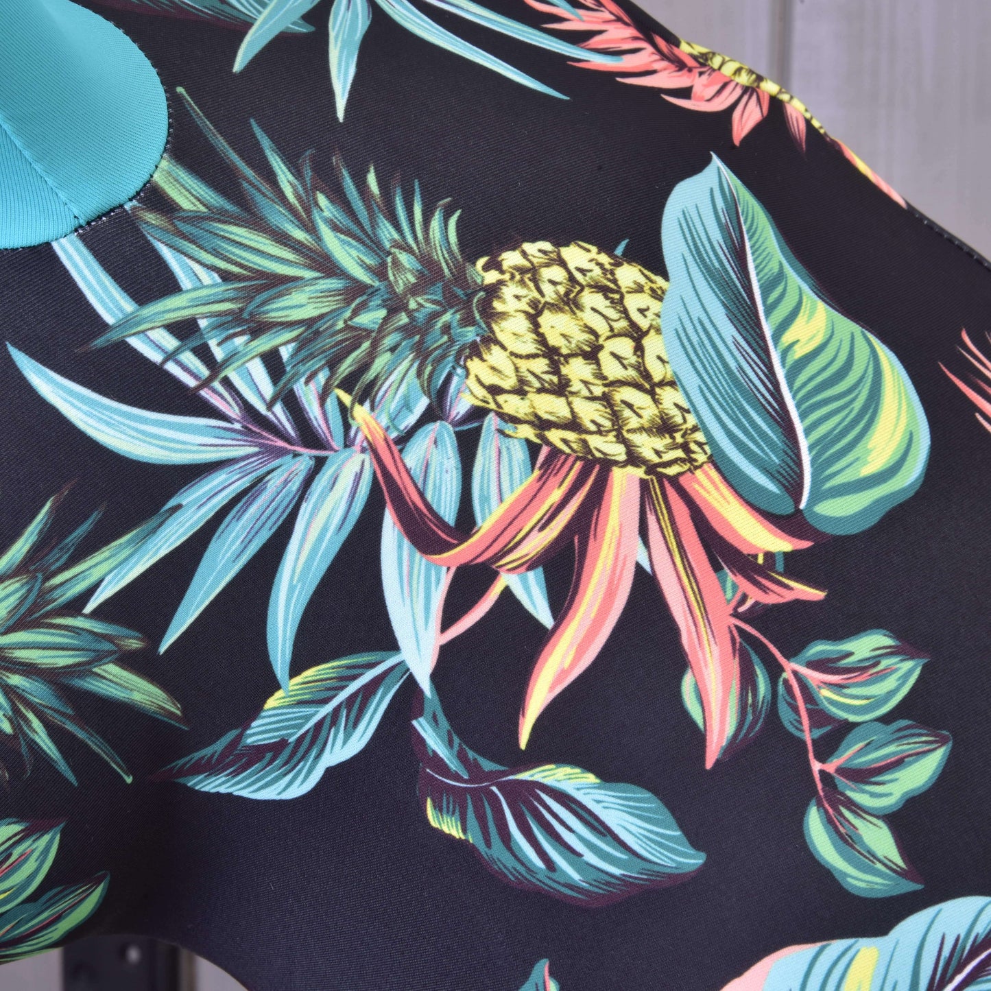 Detail Hobby Horse ochranný oblek Pineapple (Lycra) od Hobby Horse LarDen, vyrobeného z PES a Lycra, s výraznými ananasy, zelenými listy a tropickým porostem na černém podkladu, ideální pro vytvoření stylového ochranného obleku.