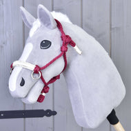 Šedý plyšový Hobby Horse od Hobby Horse LarDen s bílou lysinou, černýma knoflíkovýma očima a doplňkem Parelka Czi-Czi, na dřevěné tyči před šedou dřevěnou stěnou.