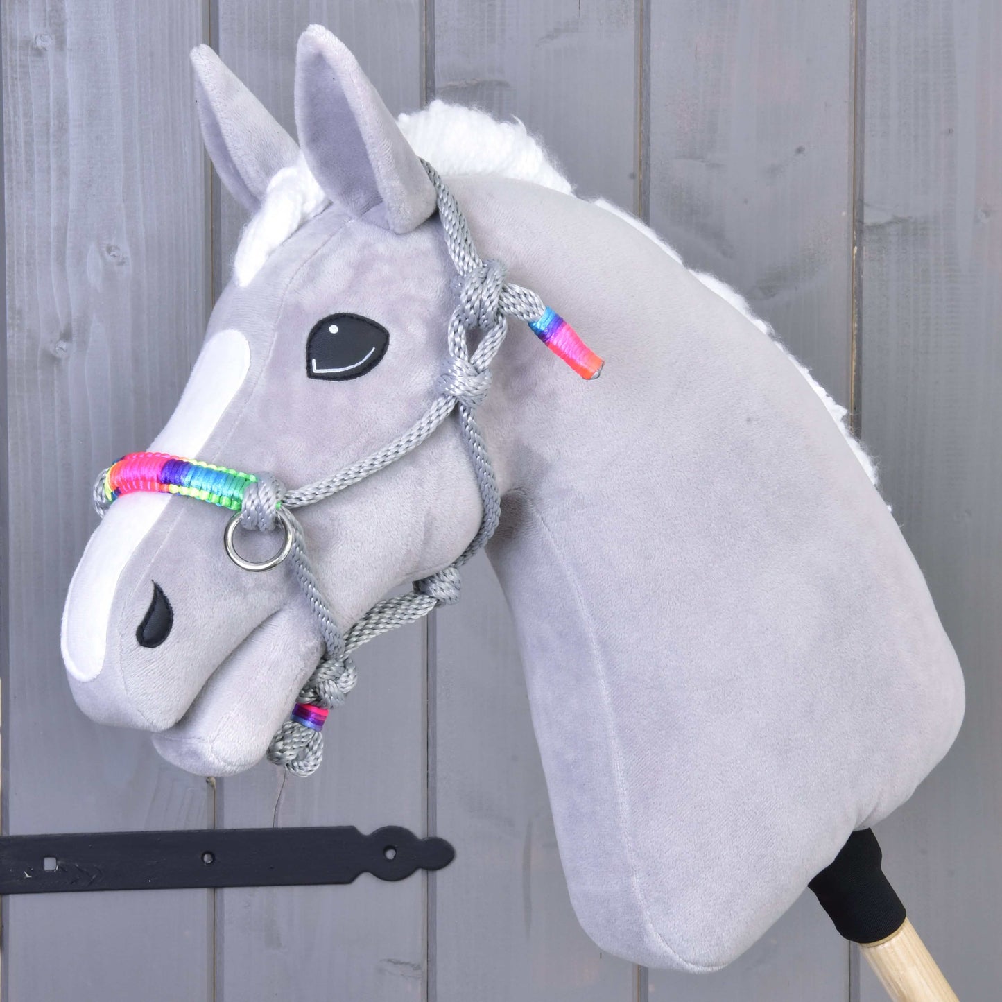 Hobby Horse LarDen Parelka Manhattan na měkkém šedém doplňku pro Hobby Horse s bílou lysinou, černýma knoflíkovýma očima a pletenou provazovou ohlávkou s duhovými akcenty, na šedém dřevěném pozadí.