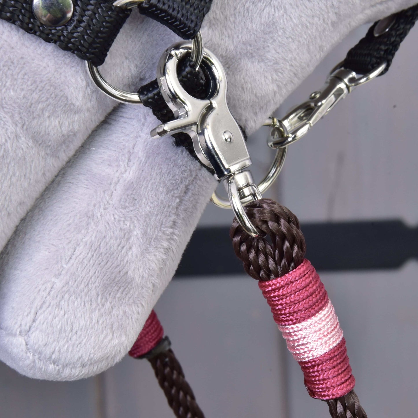 Detail kovového klipu připevňujícího Hobby Horse LarDen Otěže York — hnědé a růžové pletené polypropylenové lano — k černému textilnímu popruhu na měkkém světle šedém povrchu, pravděpodobně jako součást výbavy pro Hobby Horse.