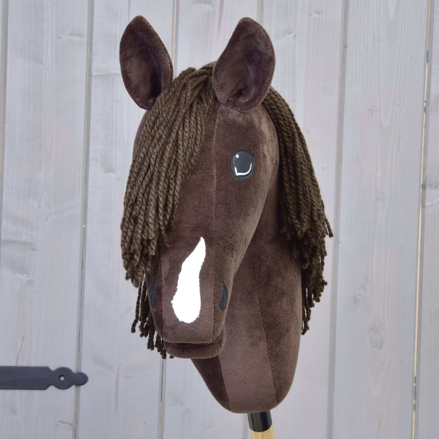 Hobby Horse Chianti s černou ohlávkou od Hobby Horse LarDen je hnědý plyšový Hobby Horse s bílou lysinou, černýma knoflíkovýma očima a hnědou přízovou hřívou, inspirovaný klidným American Quarter Horse. Na pozadí dřevěných panelů.