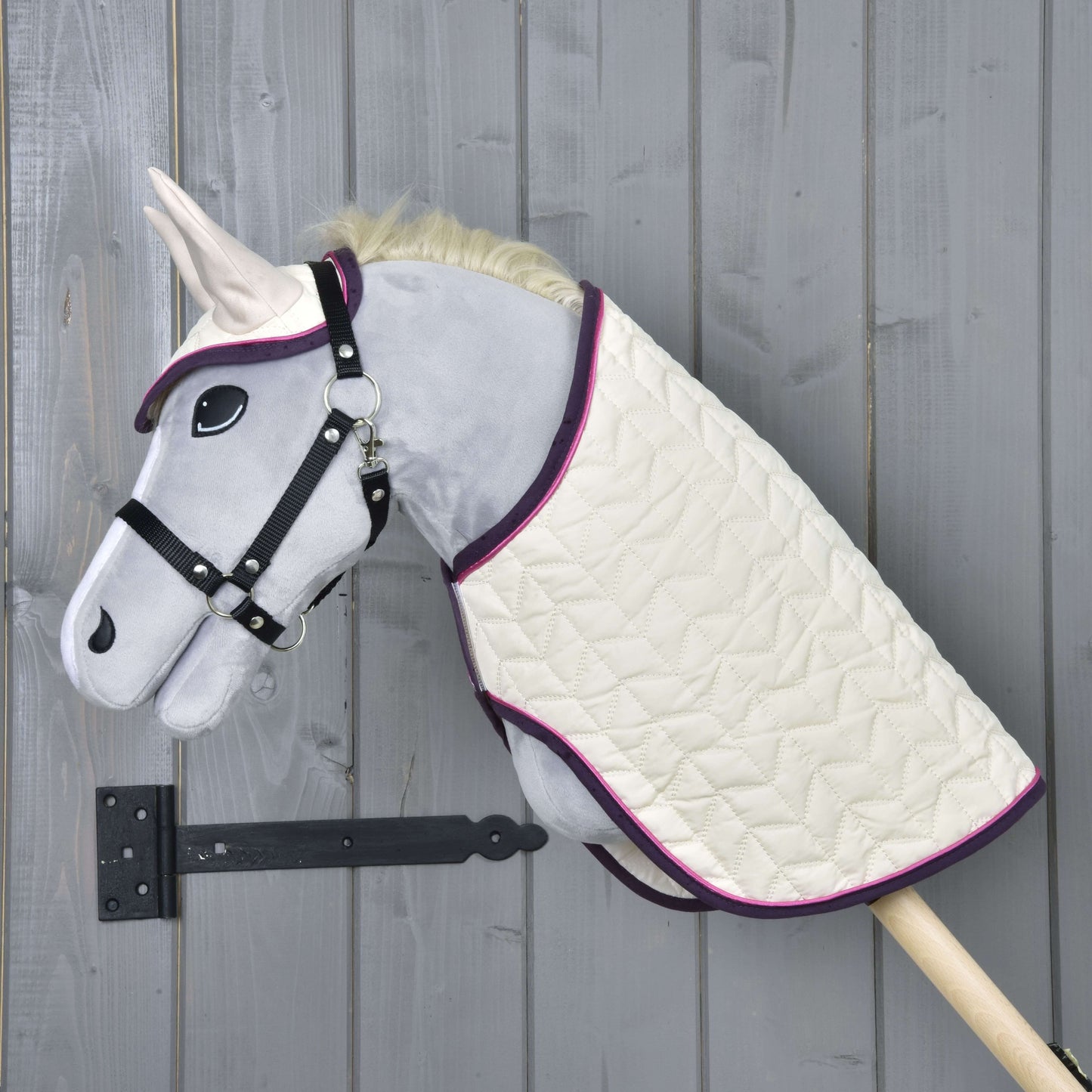Set Supreme Crema od Hobby Horse LarDen s šedým Hobby Horse, černou uzdečkou, krémovou prošívanou dekou s fialovým lemem a ladící čabrakou, nasazený na dřevěné tyči před šedou dřevěnou stěnou.