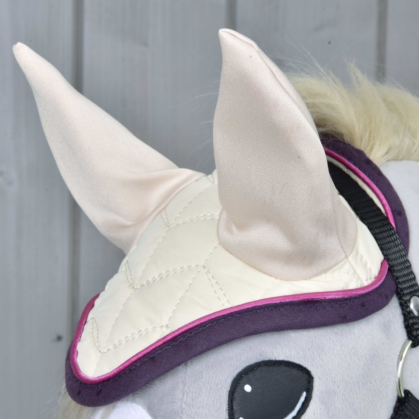 Detail Set Supreme Crema od Hobby Horse LarDen: hlava koně má krémově zbarvenou prošívanou čabraku s velkými látkovými ušimi a fialovým lemem, na pozadí světle šedých dřevěných panelů.