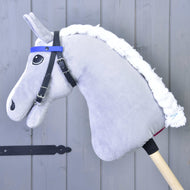 Uzdečka bez nánosníku modrá se 2 čelenkami (vel. M) od Hobby Horse LarDen na plyšovém Hobby Horse s bílou přízovou hřívou, připevněném na dřevěné tyči a opřeném o šedou dřevěnou stěnu.