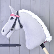 Uzdečka bez nánosníku růžová se 2 čelenkami (vel. M) od Hobby Horse LarDen na šedém Hobby Horse s bílou přízovou hřívou, černou uzdečkou bez nánosníku a růžovou čelenkou, na dřevěné tyči před světle šedými dřevěnými panely.