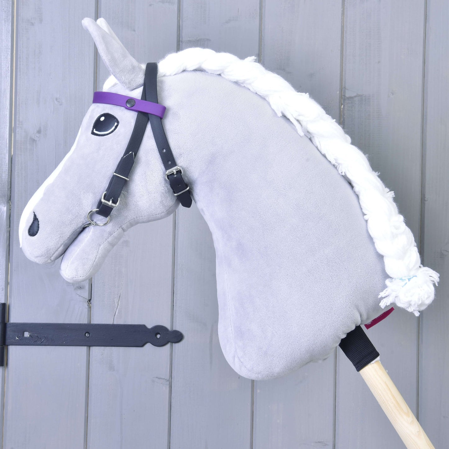 Šedý Hobby Horse s bílou přízovou hřívou a uzdečkou Hobby Horse LarDen se 2 fialovými čelenkami (vel. M), bez nánosníku, na dřevěné tyči před světle šedou dřevěnou stěnou.