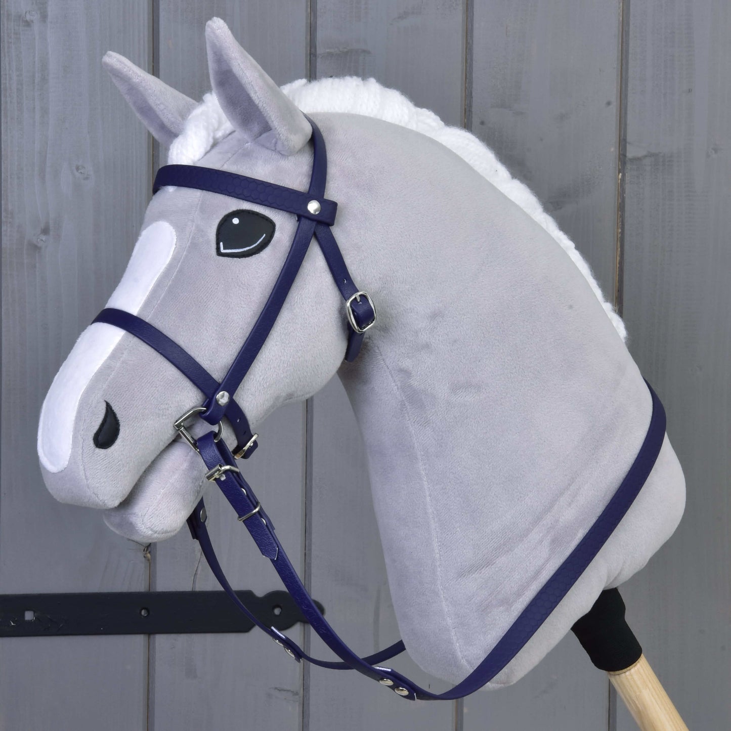 Plyšový šedý Hobby Horse od Hobby Horse LarDen s kompletní uzdečkou s nánosníkem v tmavě fialové barvě, černo-bílýma očima, bílou hřívou, nasazený na dřevěné tyči, stojí před šedou dřevěnou stěnou.
