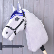 Plyšový Hobby Horse od Hobby Horse LarDen s bílou hřívou, černou uzdečkou a čabrakou DeLuxe Blue pro ochranu proti hmyzu, nasazený na dřevěné tyči před světle šedou dřevěnou stěnou.