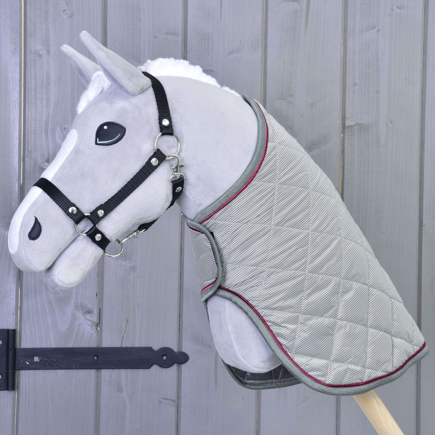 Šedý plyšový Hobby Horse od Hobby Horse LarDen s černou uzdečkou a dekou Silver Eleganza stojí před dřevěnou stěnou se svislými panely.