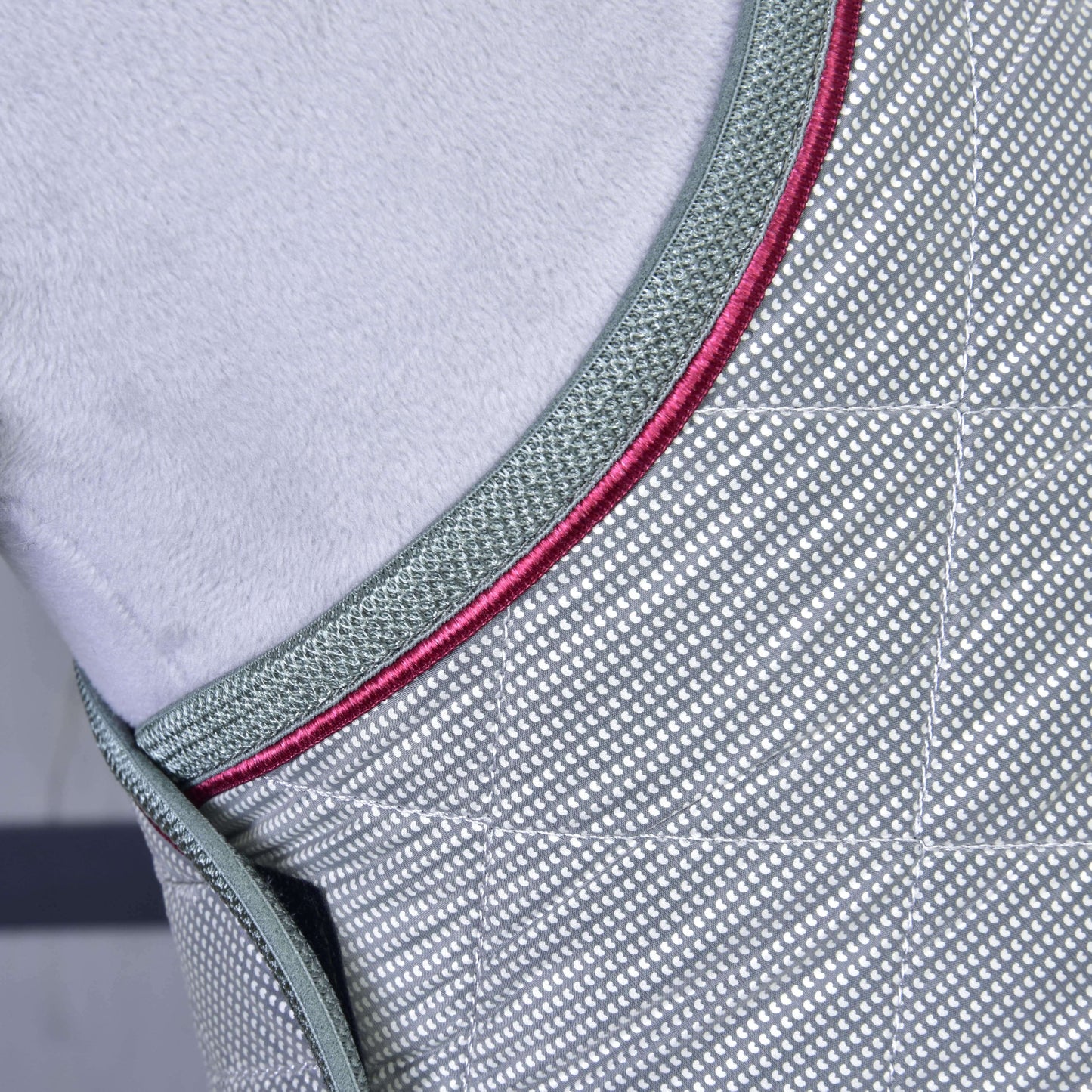 Detail deky Silver Eleganza od Hobby Horse LarDen: šedá tréninková deka s kosočtvercovým vzorem, tmavě zeleným lemováním a červenými akcenty. Měkké světle šedé pozadí zvýrazňuje její detailní prošívání pro optimální ochranu koně.
