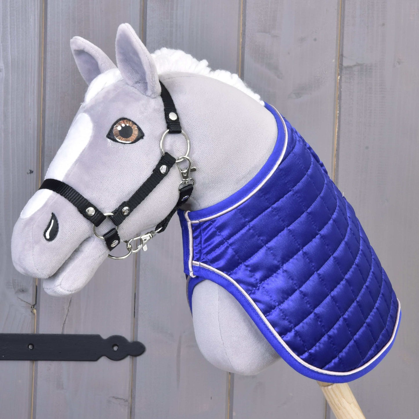 Plyšový Hobby Horse s bílou hřívou, černou ohlávkou a dekou Hobby Horse LarDen Deka DeLuxe Blue oboustranná pro extra ochranu, nasazený na dřevěné tyči na pozadí šedého dřeva.