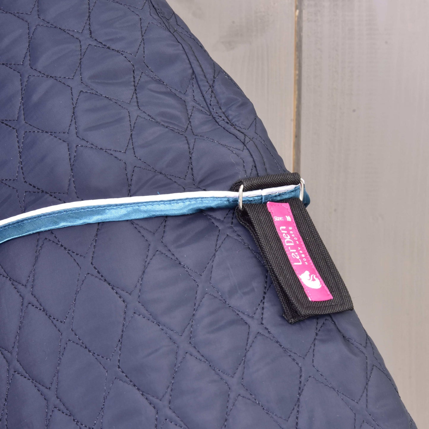 Deka s odepínatelným krkem Mandarin od Hobby Horse LarDen v tmavě modré, detail se světle modrým páskem s kovovým kroužkem a černo-růžovým štítkem s textem a grafikou koně. Na světlém dřevěném pozadí.
