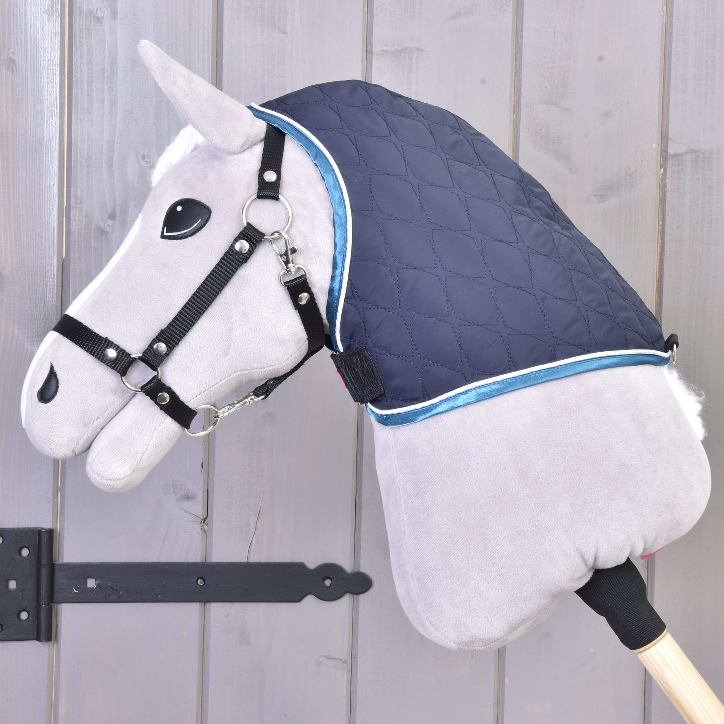 Plyšový Hobby Horse LarDen s černou ohlávkou a dekou s odepínatelným krkem Mandarin, nasazený na dřevěné tyči, zobrazený před dřevěnou stěnou.