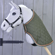 Šedý plyšový Hobby Horse na tyči s Dekou Sport od Hobby Horse LarDen v zelené barvě s oranžovým lemováním a černou ohlávkou stojí před dřevěnou stěnou s černým železným závěsem.