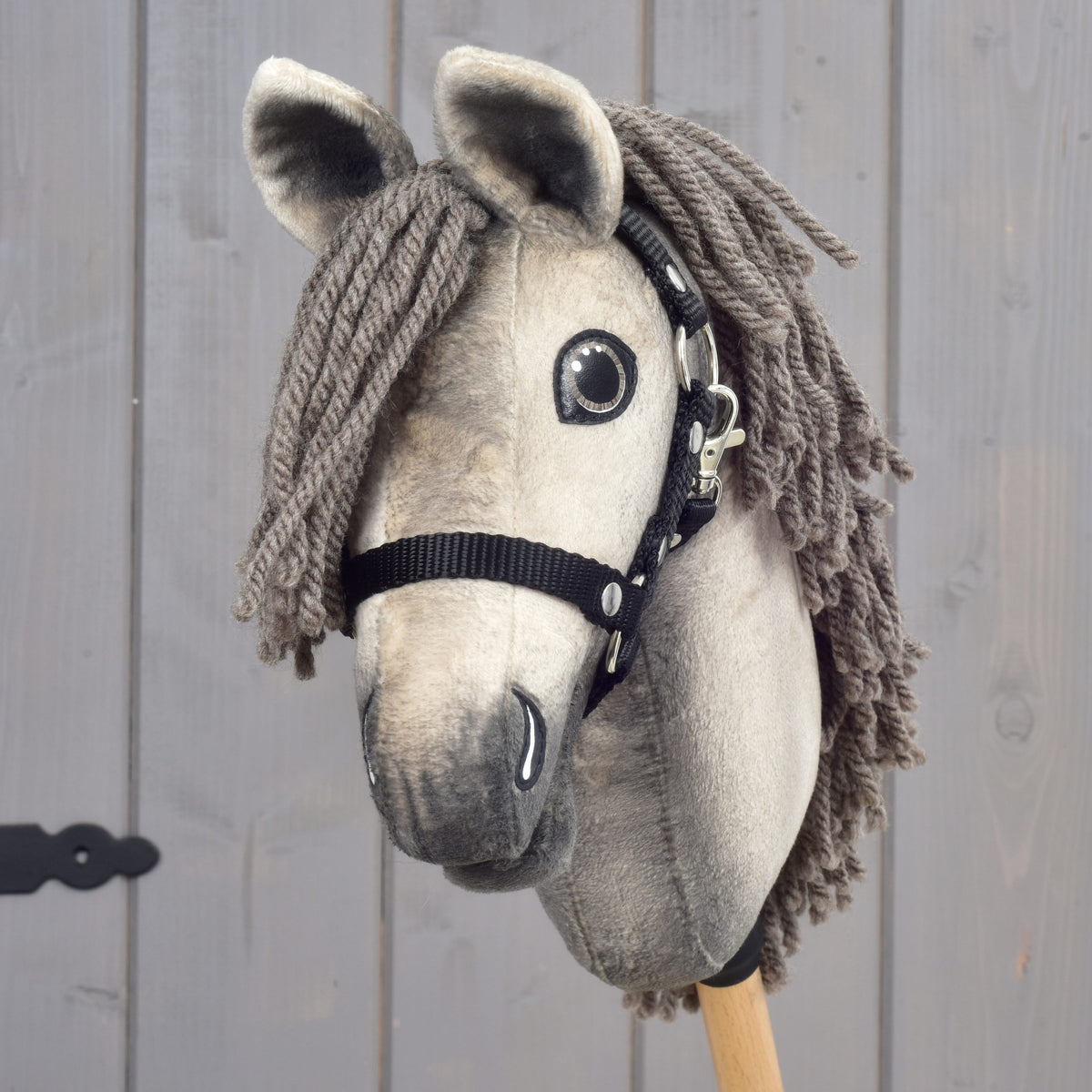 Detailní záběr na Hobby Horse Dusty s černou ohlávkou ve velikosti XS od Hobby Horse LarDen podle hannoverského hřebce, se šedou přízovou hřívou, černou ohlávkou a výraznýma knoflíkovýma očima, nasazený na dřevěné tyči na pozadí šedého dřeva.