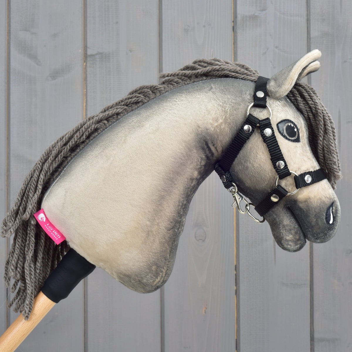 Hobby Horse Dusty s černou ohlávkou ve velikosti XS od Hobby Horse LarDen má realistickou šedou plyšovou hlavu hannoverského koně s knoflíkovýma očima a černou ohlávkou a je nasazený na dřevěné tyči. Zobrazený je na pozadí dřevěných prken.