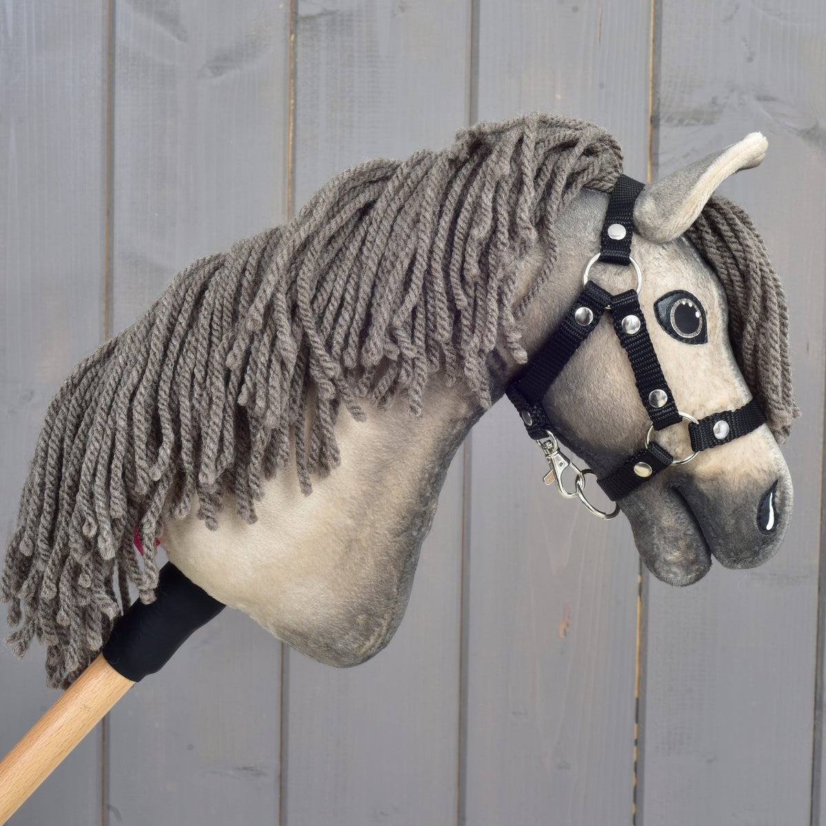 Hobby Horse Dusty s černou ohlávkou ve velikosti XS od Hobby Horse LarDen se šedou přízovou hřívou a dřevěnou tyčí jako držadlem je zobrazený před šedým dřevěným plotem.