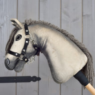 Hobby Horse Dusty s černou ohlávkou ve velikosti XS od Hobby Horse LarDen má plyšovou hlavu hannoverského koně s knoflíkovýma očima a černou ohlávkou, je nasazený na dřevěné tyči a zobrazený na pozadí světlého dřevěného panelu.