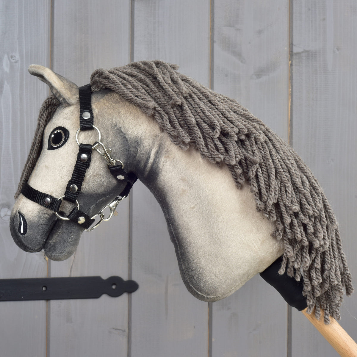 Hobby Horse Dusty s černou ohlávkou ve velikosti XS od Hobby Horse LarDen se šedou srstí, přízovou hřívou a černou ohlávkou je nasazený na dřevěné tyči a umístěný před světle šedou dřevěnou stěnou.