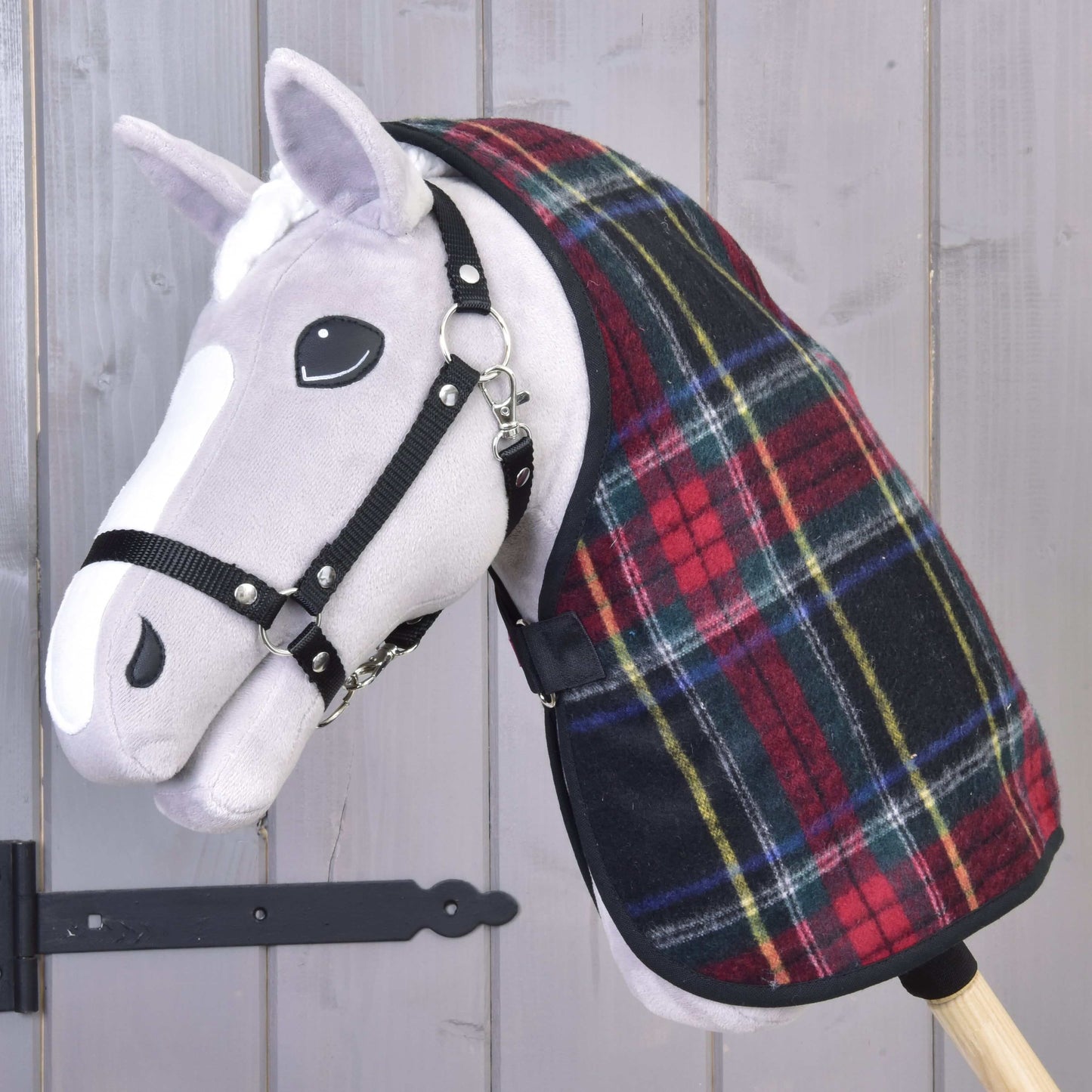 Plyšový Hobby Horse s bílým obličejem a černou uzdečkou má na sobě deku Deka stájová Edinburgh od Hobby Horse LarDen v červené a zelené kostce, pózuje před šedou dřevěnou stěnou s viditelným černým kovovým pantem.