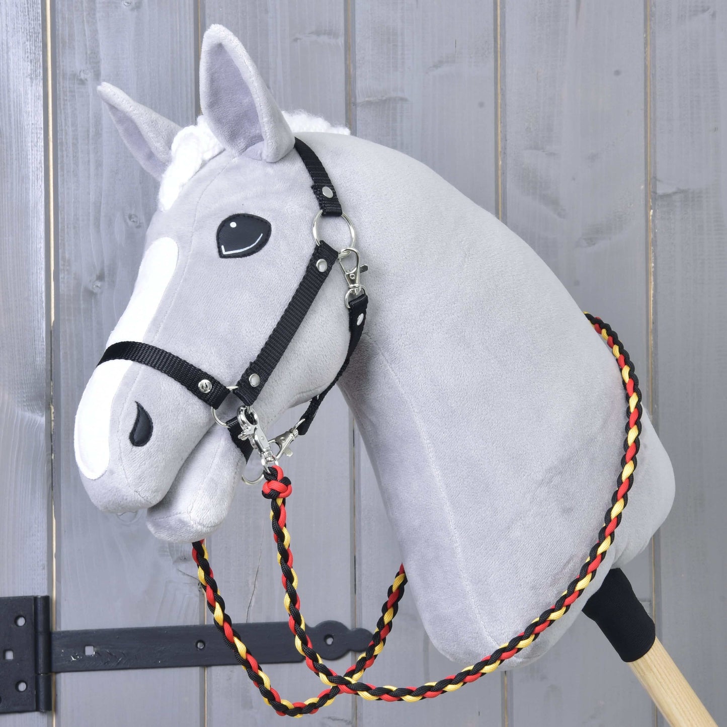 Hobby Horse LarDen Otěže Flag na šedém plyšovém Hobby Horse na dřevěné tyči, s černým postrojem a ručně vyráběnými otěžemi z pleteného červeného, žlutého a černého polypropylenového lana, před dřevěnou stěnou.