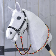 Hobby Horse LarDen Otěže Flag na šedém plyšovém Hobby Horse na dřevěné tyči, s černým postrojem a ručně vyráběnými otěžemi z pleteného červeného, žlutého a černého polypropylenového lana, před dřevěnou stěnou.