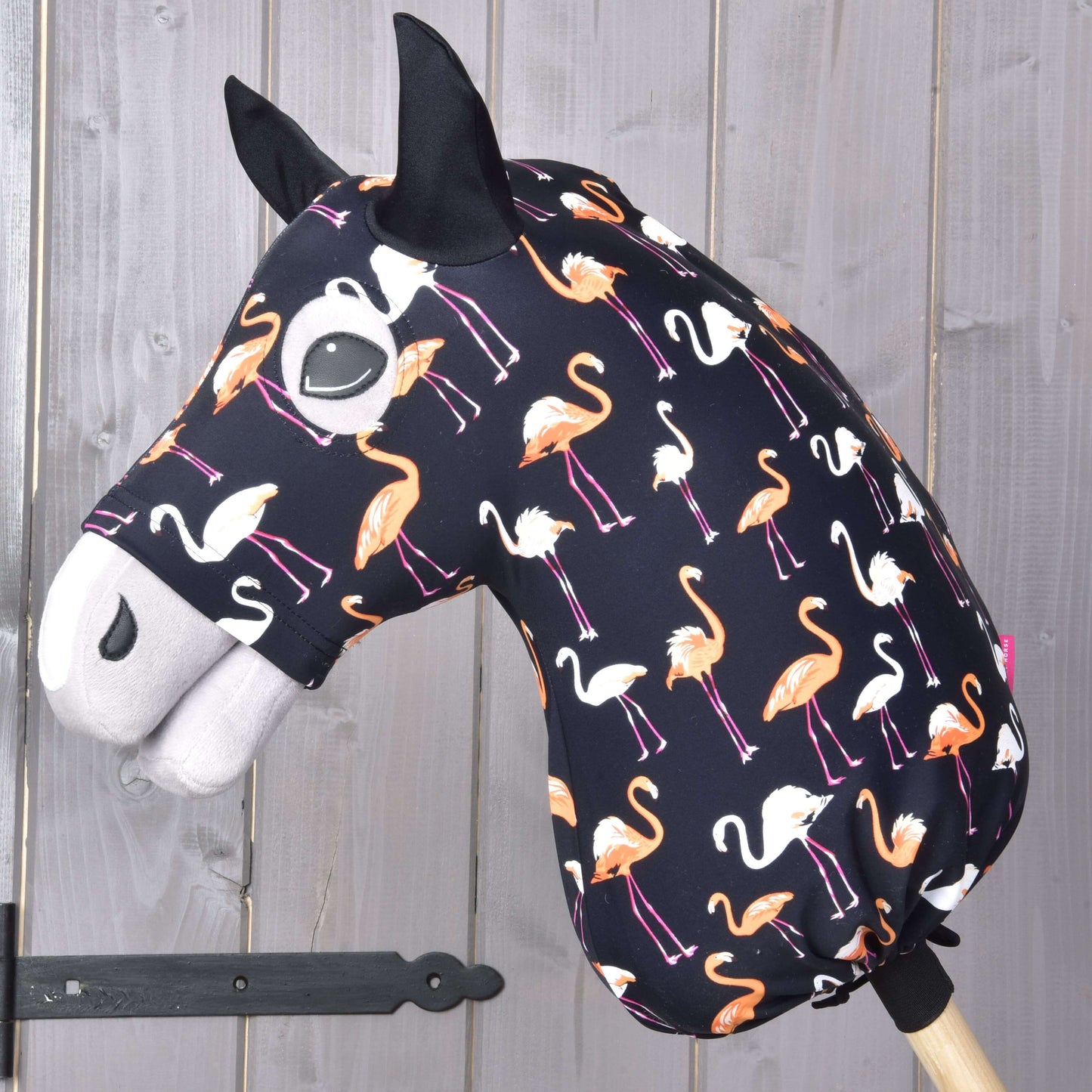 Hobby Horse Lycra ochranný oblek Flamingo od Hobby Horse LarDen s lycrovým potahem s oranžovými a bílými plameňáky na tmavém podkladu, nasazený na dřevěné tyči před stěnou z dřevěných panelů.
