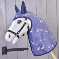 Pláštěnka Flamingo od Hobby Horse LarDen, šedý Hobby Horse v tmavě modré pláštěnkové kapuci s růžovými plameňáky, je umístěna před dřevěnou stěnou s černým kovovým pantem.
