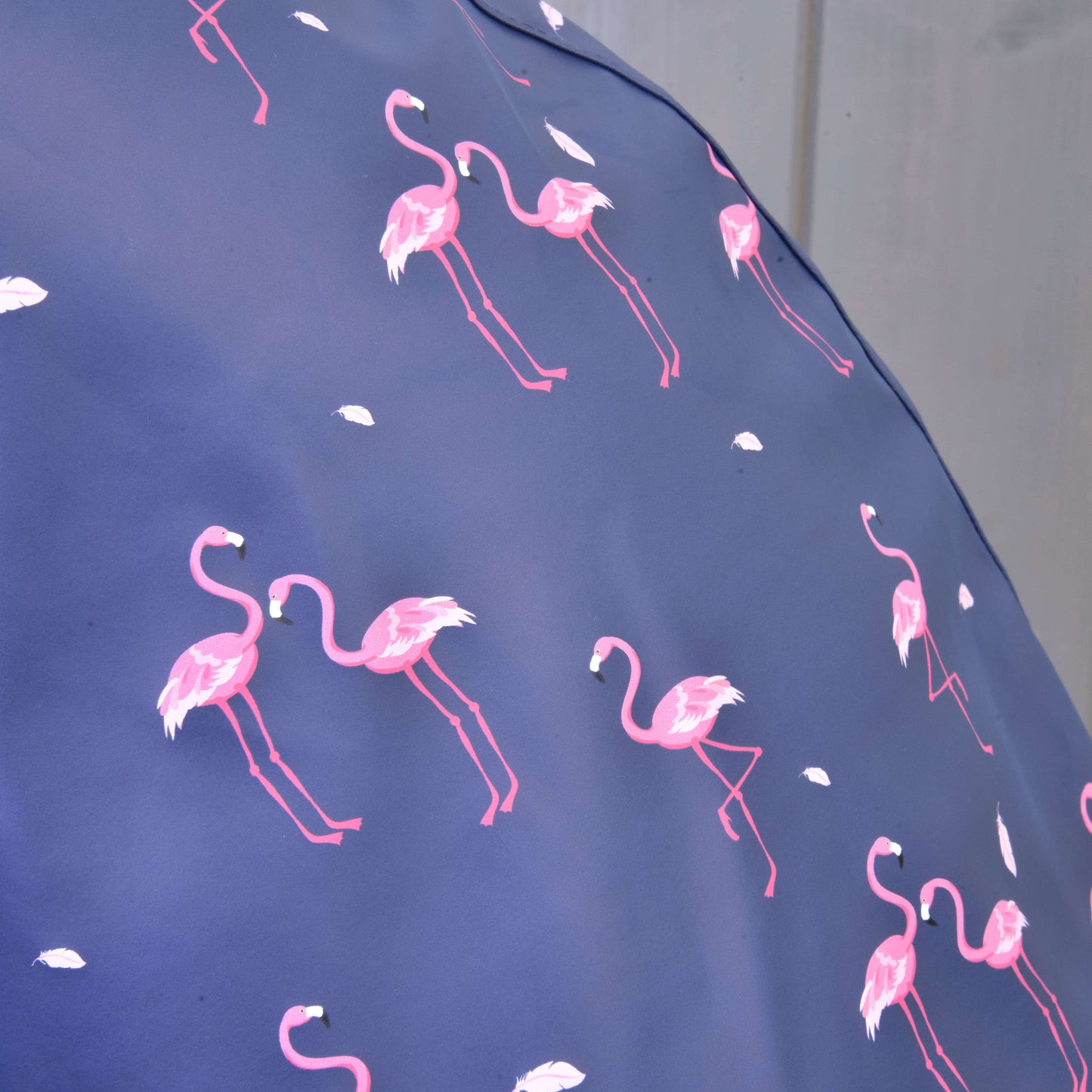 Detail Pláštěnky Flamingo od Hobby Horse LarDen ukazuje modrou látku s růžovými plameňáky a bílými pery na jemně rozostřeném pozadí – ideální do každého počasí nebo pro hravé dny.