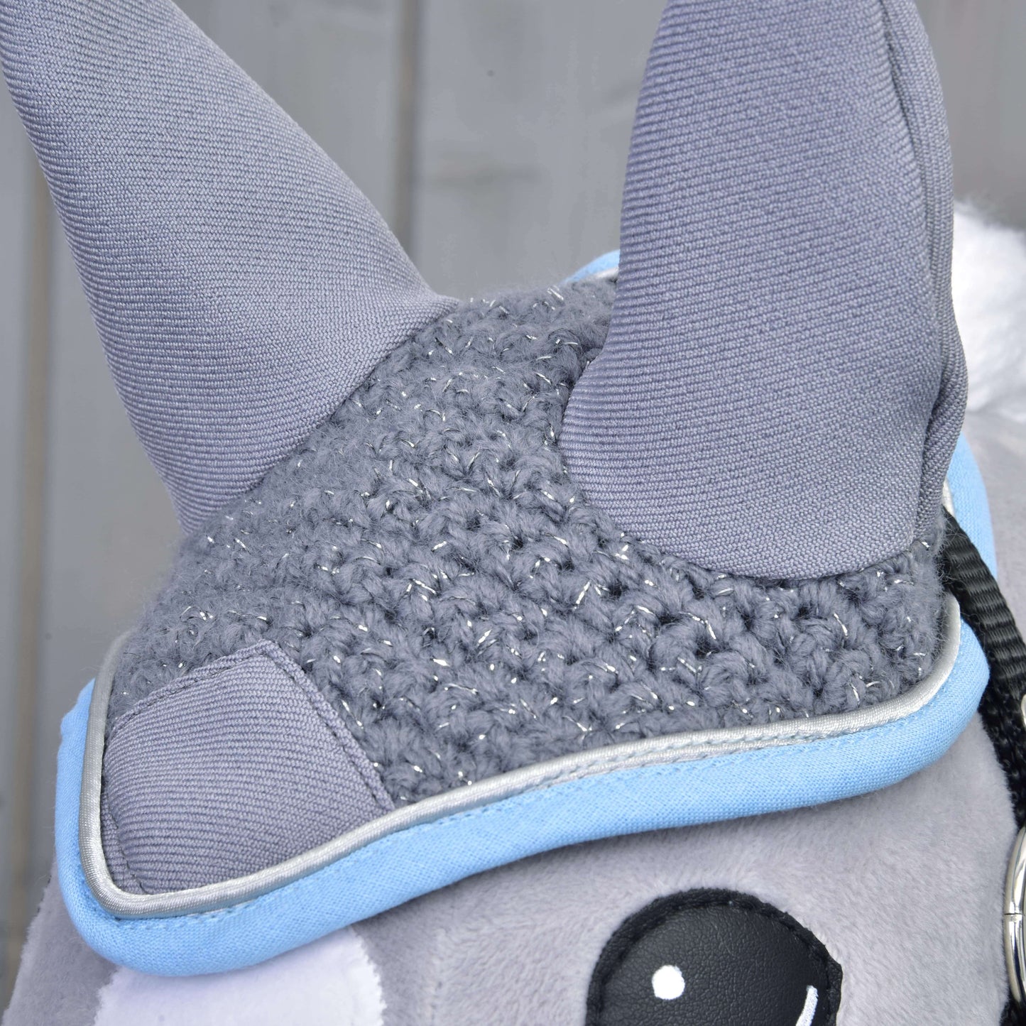 Detail plyšové hračky Hobby Horse LarDen s čabrakou „Čabraka háčkovaná šedá“, se dvěma šedými háčkovanými ušima, pletenou šedou částí se stříbrnými nitěmi, světle modrým lemem a částí černobílého oka. Rozmazané pozadí.