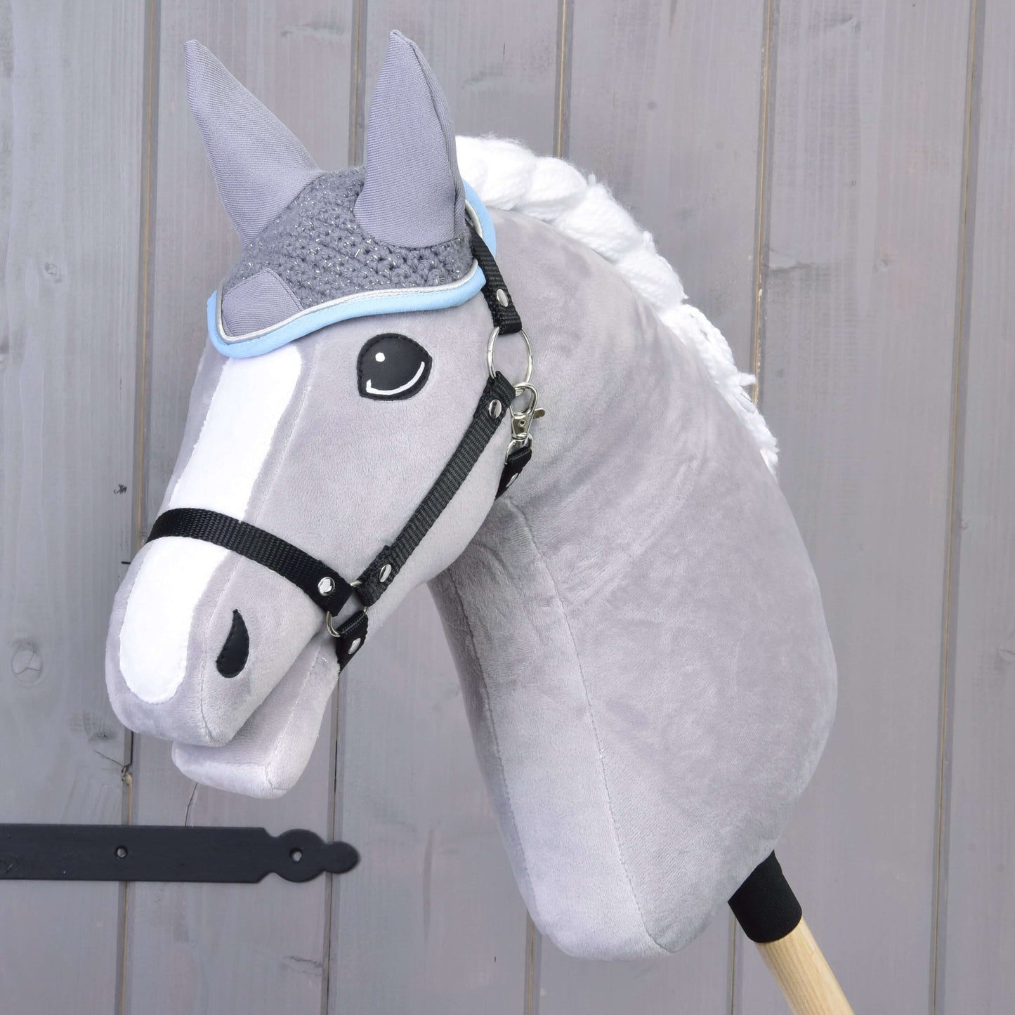 Čabraka háčkovaná šedá od Hobby Horse LarDen na plyšovém šedém Hobby Horse s bílou hřívou a černou uzdou, nasazeném na dřevěné tyči před světlou dřevěnou stěnou.