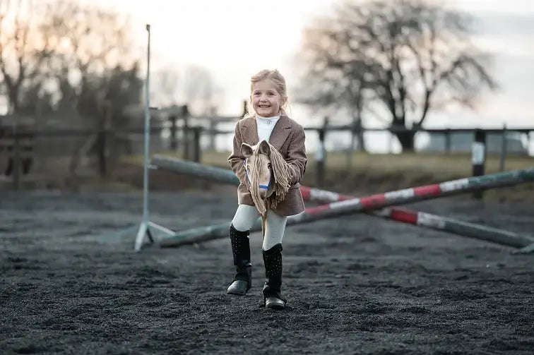 Malá dívka se usmívá při hobby horsing, venku na hliněné jízdárně předstírá jízdu na hračce hobby horse, v pozadí jsou zkřížené tyče a stromy. Má na sobě hnědou bundu, bílé kalhoty a černé jezdecké boty.