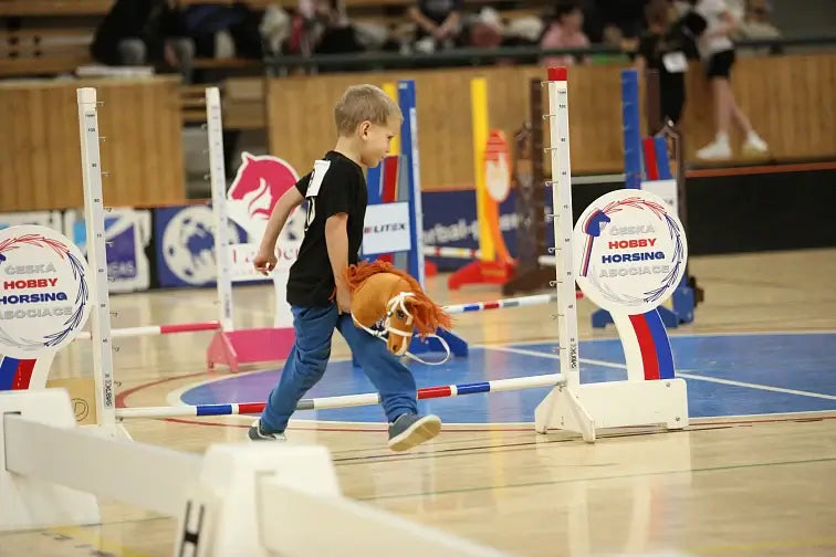 Malý chlapec se účastní soutěže Hobby Horse, jde s hračkovým koněm po podlaze tělocvičny a kolem něj jsou barevné skokové překážky a cedule akce Hobby Horsing.