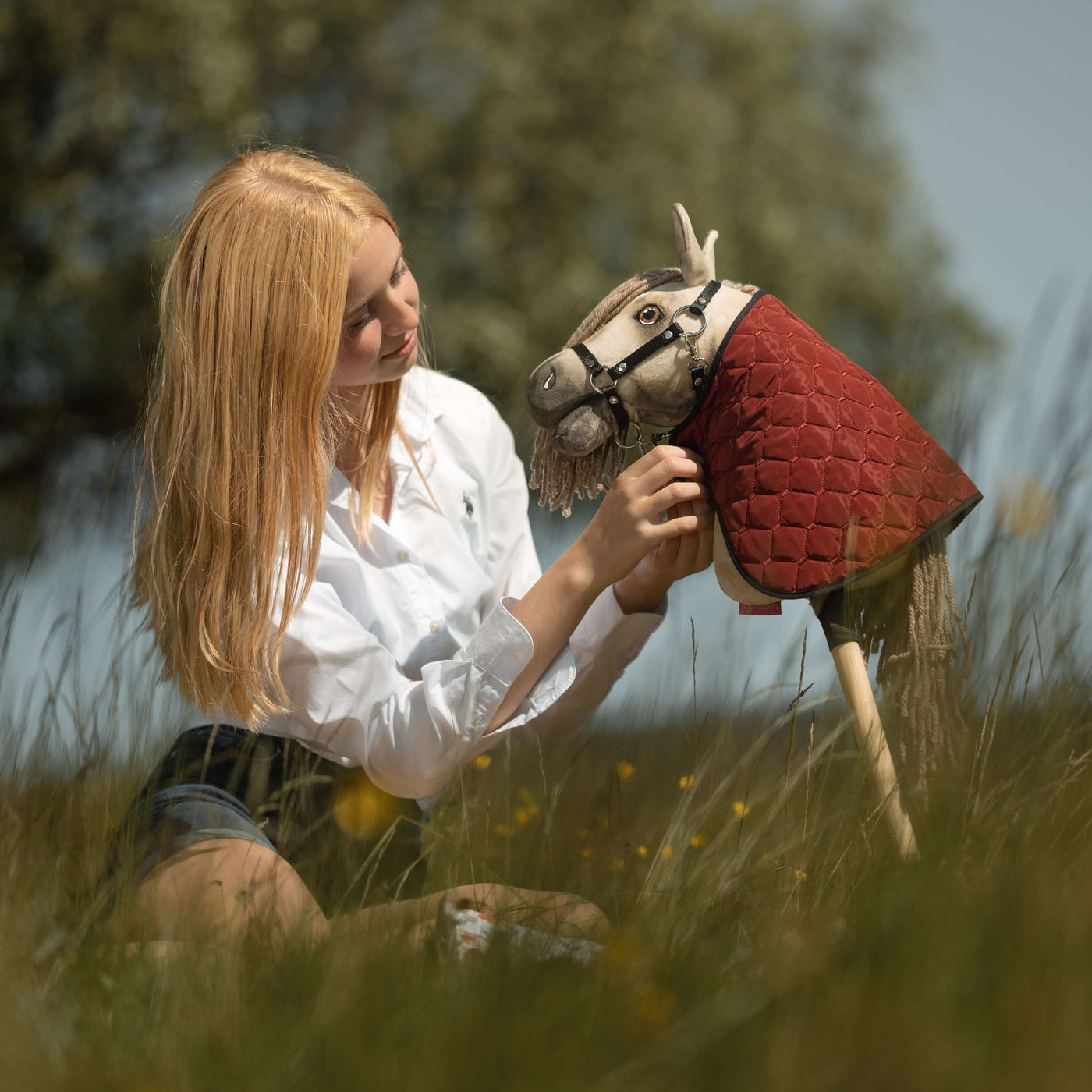 Mladá žena s blond vlasy sedí ve vysoké trávě, usmívá se a drží svého společníka pro Hobby Horsing – Hobby Horse oblečeného do červeného prošívaného pláště. Pozadí tvoří stromy a rozostřená zeleň.